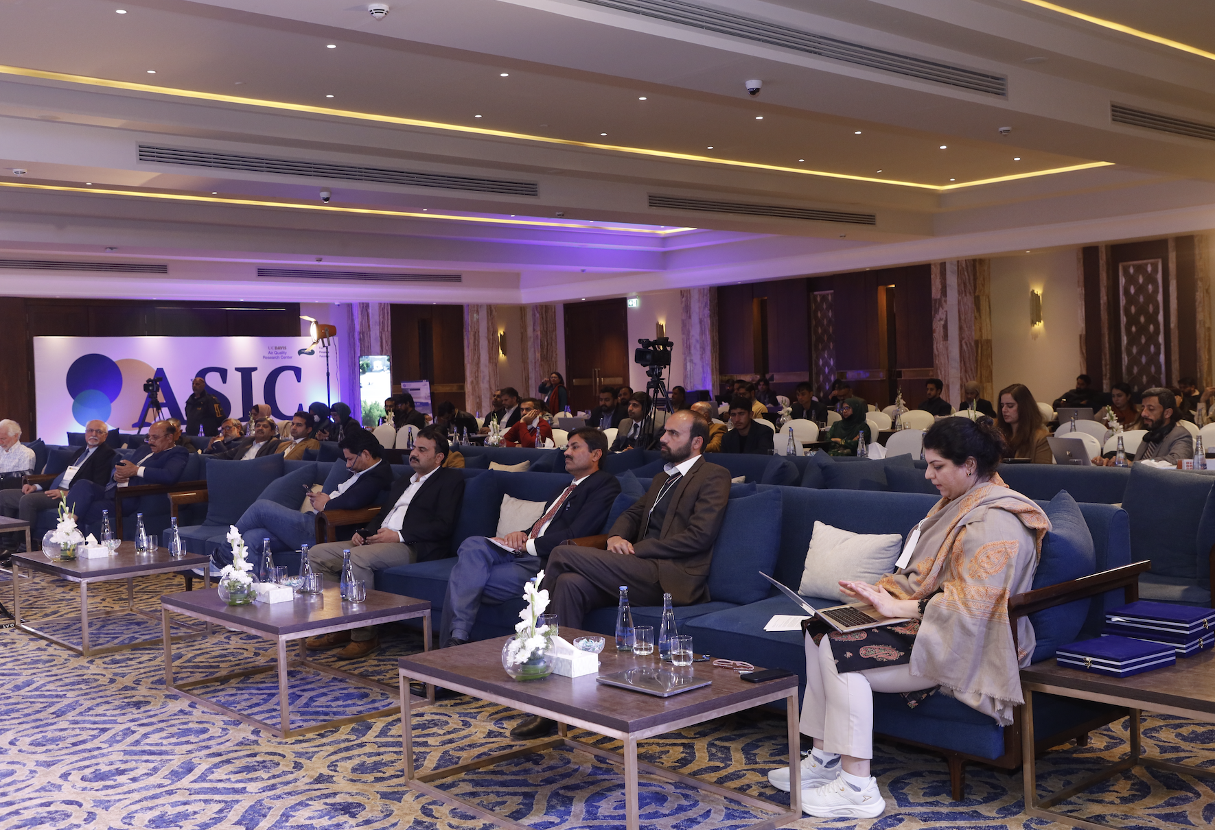 Audience seated in a conference hall, facing a presentation stage with a large sign.
