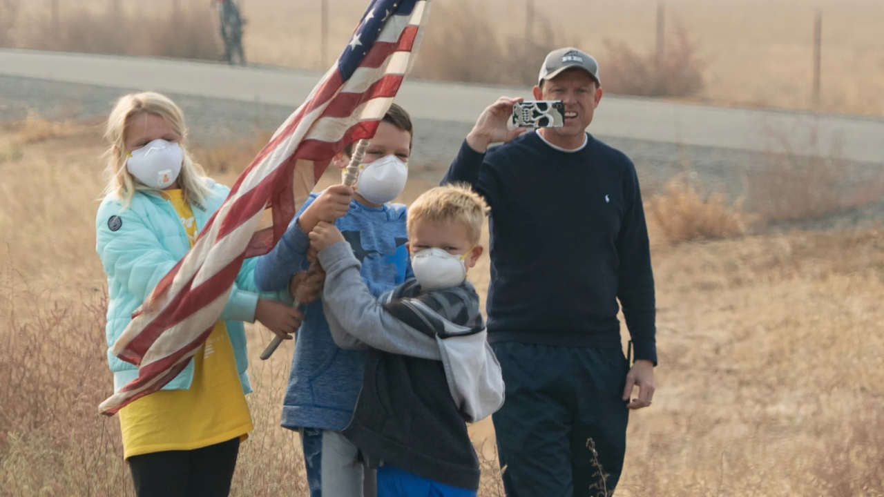 Image: people outside wear face masks and wave an American flag.