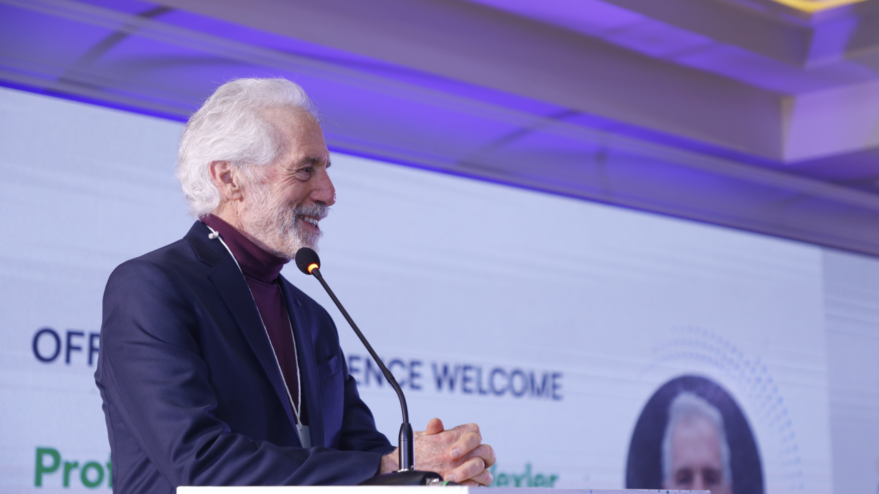 A man with gray hair speaks at a podium during a conference, welcome banner in the background.